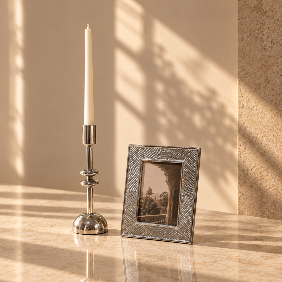A silver-toned candlestick holder with a white taper candle stands beside a textured silver picture frame on a warm beige marble surface, with soft window shadow patterns cast across the background.