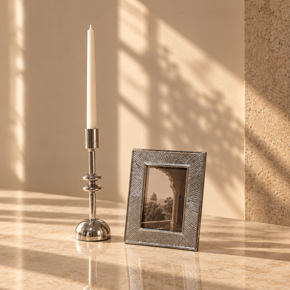 A silver-toned candlestick holder with a white taper candle stands beside a textured silver picture frame on a warm beige marble surface, with soft window shadow patterns cast across the background.