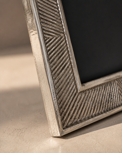 Close-up of a textured silver picture frame corner with a geometric herringbone pattern, resting on a beige surface with a dark background.