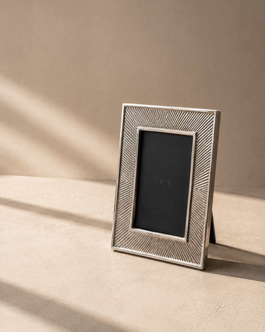 A rectangular silver picture frame with a textured, geometric chevron pattern border, standing on a beige surface against a matching beige background with soft diagonal shadows.