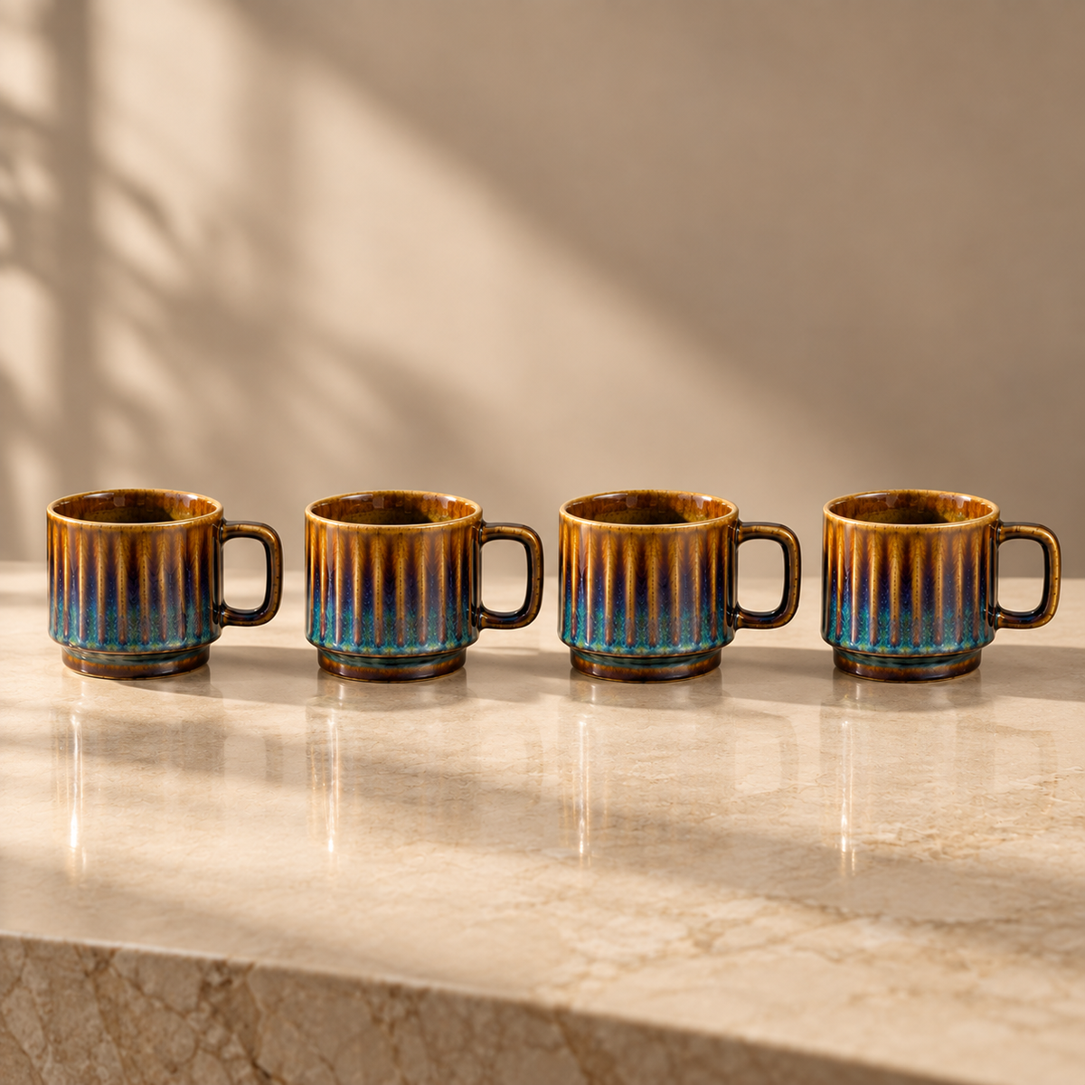 Four small ceramic cups with ribbed sides and dark handles, glazed in a warm brown and blue gradient, lined up on a light marble countertop with soft shadow projections on the wall.