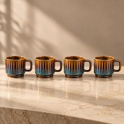 Four small ceramic cups with ribbed sides and dark handles, glazed in a warm brown and blue gradient, lined up on a light marble countertop with soft shadow projections on the wall.