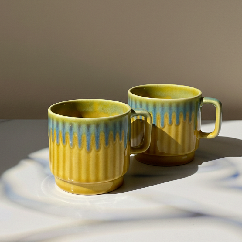 Two yellow ceramic mugs with a blue-green drip glaze and vertical ridges, sitting on a white surface with dramatic shadows against a warm beige background.