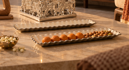 A set of decorative silver-toned serving trays arranged on a polished beige marble table, styled with fruits, nuts, and small bowls in a warm indoor setting.