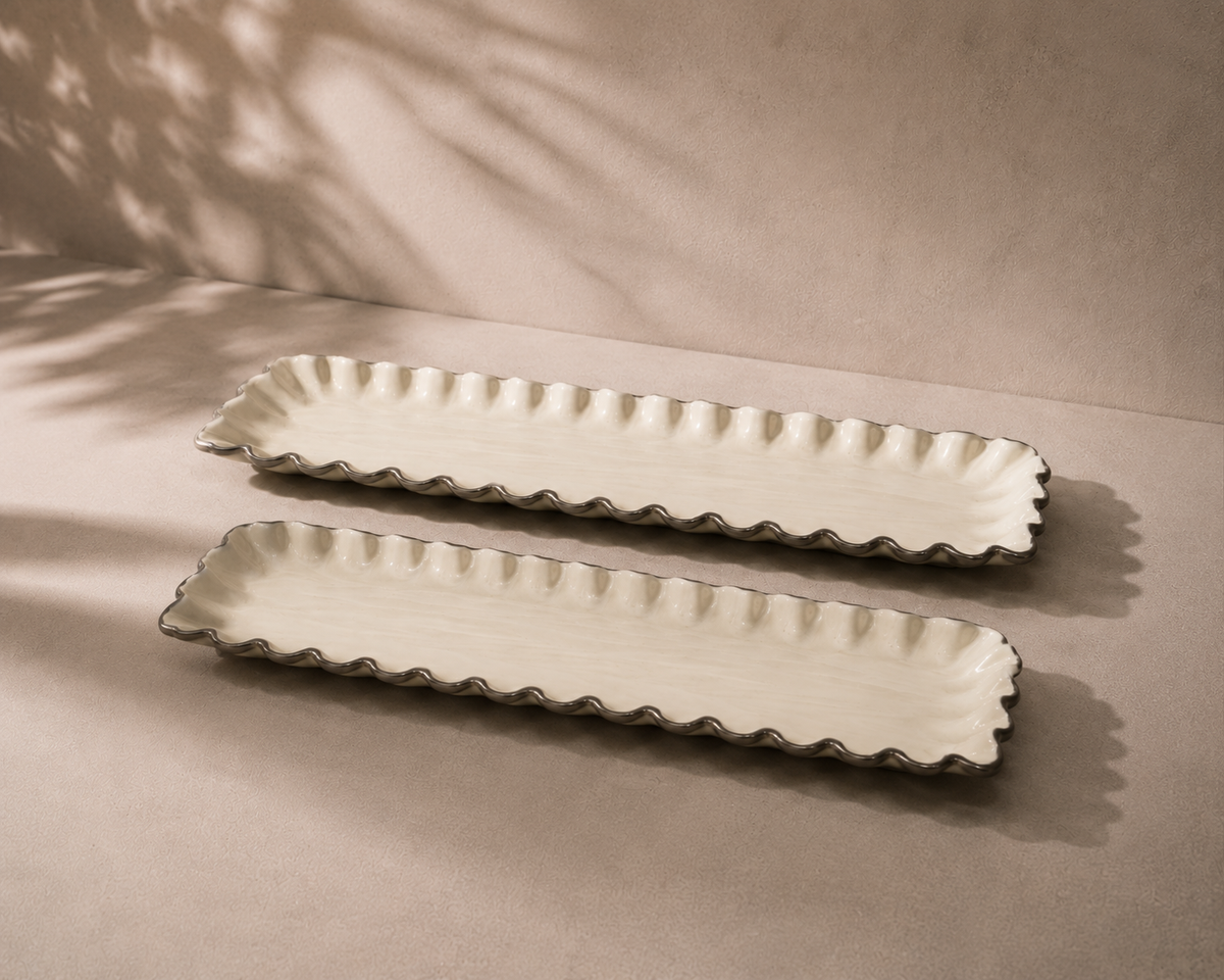 Two long rectangular ceramic serving platters with scalloped edges, shown on a neutral beige surface with soft leaf shadows cast across the scene.
