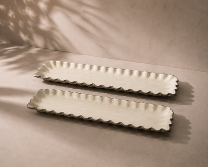 Two long rectangular ceramic serving platters with scalloped edges, shown on a neutral beige surface with soft leaf shadows cast across the scene.