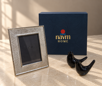 A silver textured picture frame displayed next to a navy blue 'navm HOME' gift box and two glossy black bird figurines on a beige marble surface with warm window light shadows.