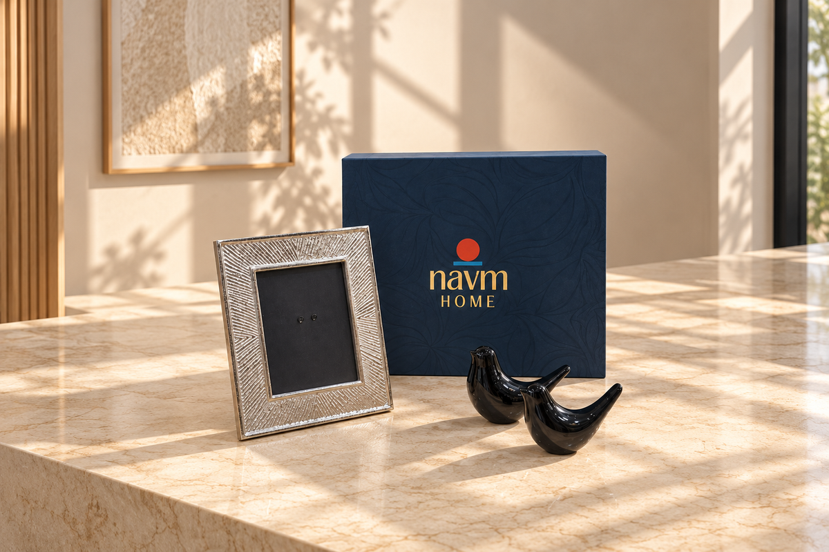 A navy blue Nav Home gift box with gold lettering stands on a beige stone surface next to a textured silver picture frame and two small black bird figurines, with warm sunlight casting shadows across the scene.