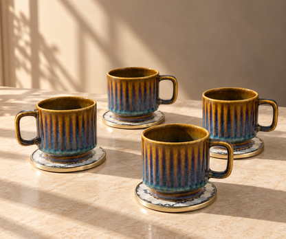 Four ceramic coffee mugs with matching saucers, featuring a textured vertical ribbed pattern in metallic gold and deep blue, arranged on a light beige stone surface with warm natural sunlight and shadows.