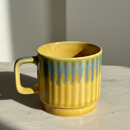 A yellow ceramic mug with a ribbed texture and a blue-green glazed drip design around the rim, sitting on a light marble surface in natural sunlight.
