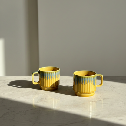 Two yellow ceramic mugs with blue-green vertical drips on the sides, placed on a light marble surface in a sunlit room.