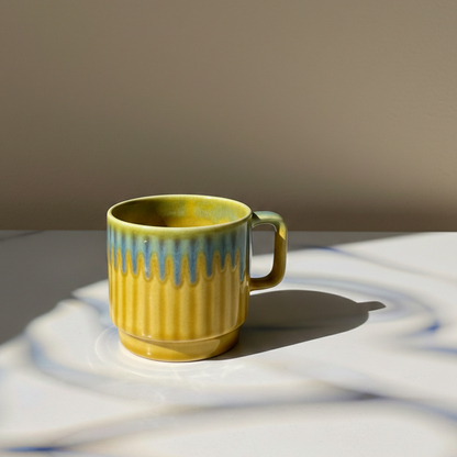 A yellow ceramic mug with a blue-green drip glaze and vertical fluting, placed on a white surface with strong sunlight and shadows.