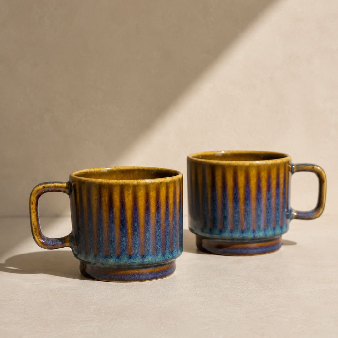 Two ceramic mugs with a ribbed texture and a glazed finish showing yellow, blue, and brown colors, set against a warm beige background with diagonal shadows. The brand logo 'navm HOME' is visible at the top.