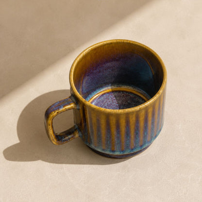 A ceramic coffee mug with a textured vertical rib design, featuring a metallic gold exterior and a deep blue-purple glazed interior, shown on a beige background with a soft shadow. The brand logo 'navm HOME' is visible at the top.